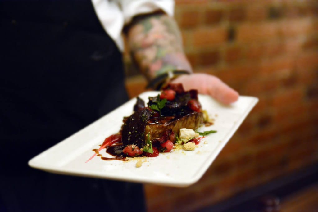 Tarjoilijan kädessä pieni gourmet-liha-annos kastikkeella ja yrteillä valkoisella lautasella.