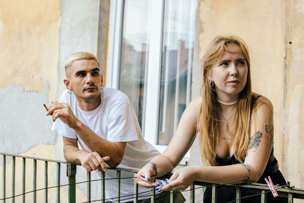 Nuori nainen ja nuori mies nojaavat parvekkeen kaiteeseen ja polttavat savukkeita.