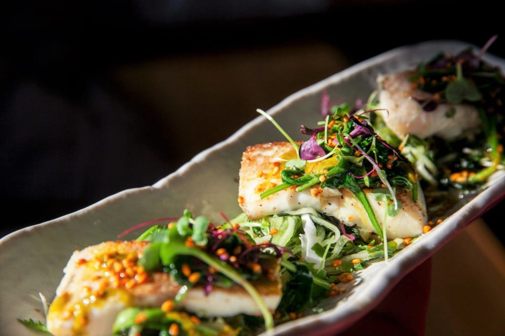 Paistettu kala-annos pitkällä lautasella, päällä vihreitä kasviksia ja siemeniä.