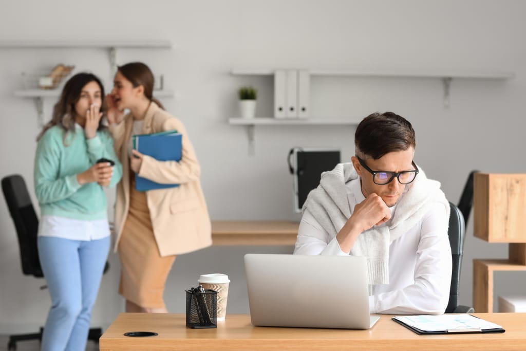 Työntekijä istuu työpöydän ääressä läppärin kanssa ja näyttää huolestuneelta, kun taustalla kaksi kollegaa kuiskuttelee keskenään ja katsoo häntä.