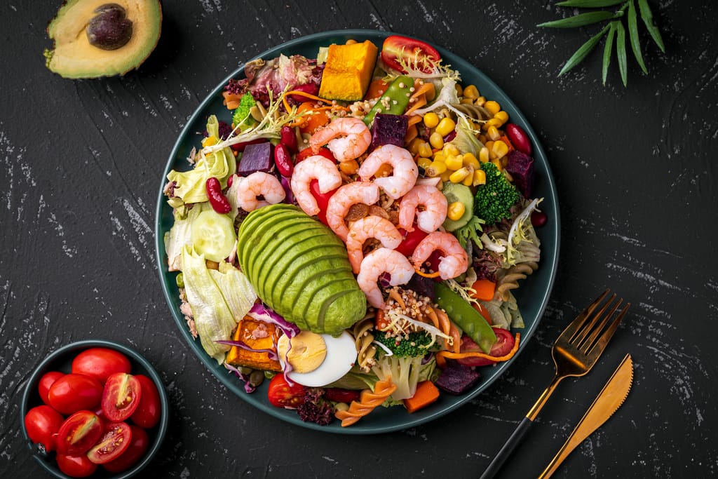 Värikäs salaattiannos kulhossa: katkarapuja, viipaloitua avokadoa, keitettyä kananmunaa, papuja, maissia, vihanneksia ja pastaa tummalla taustalla.