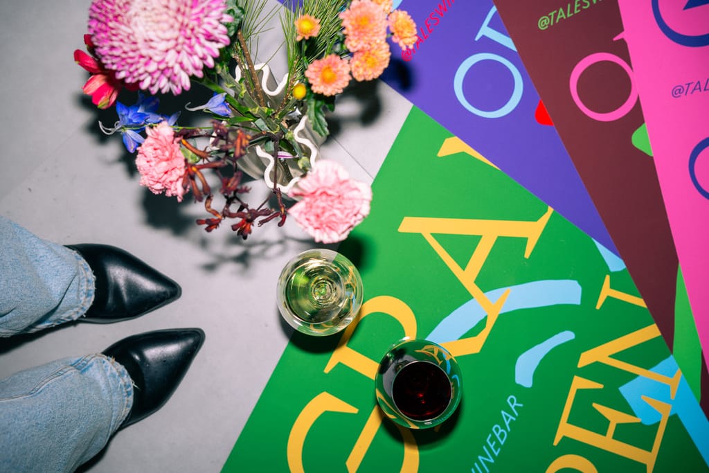 Viinilasit ja kukkakimppu värikkäiden Tales-viinibaarin julisteiden päällä.