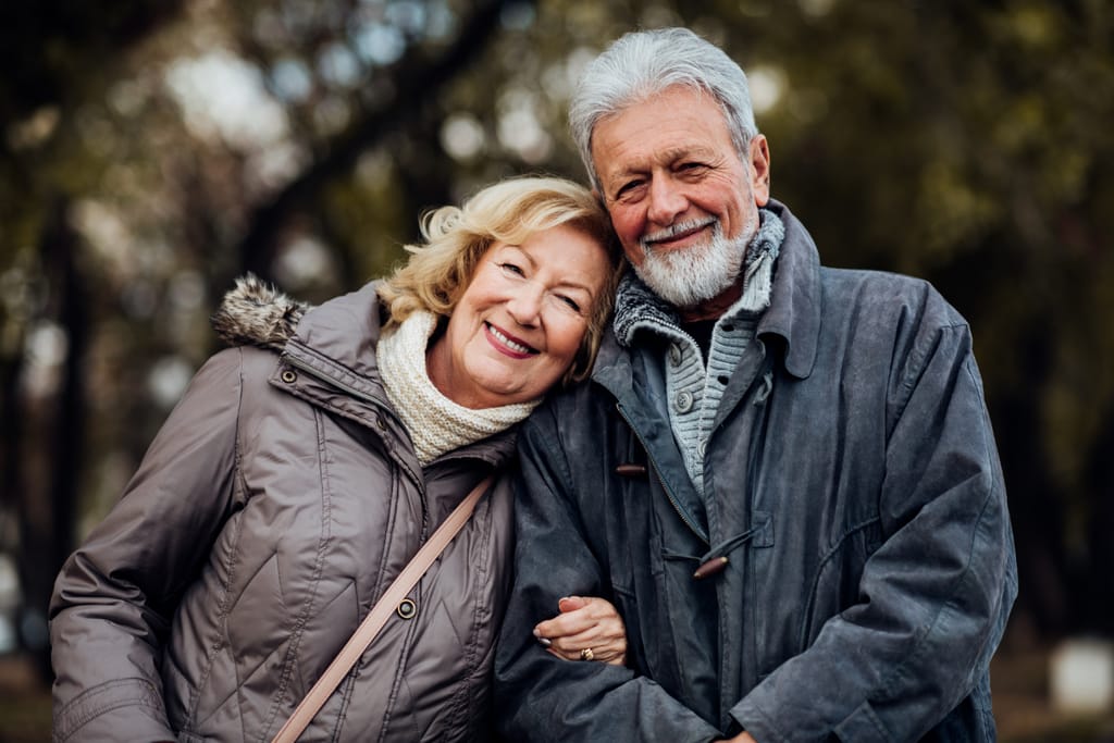 Iäkäs pariskunta seisoo ulkona puistomaisessa ympäristössä. Nainen nojaa hymyillen miehen olkapäähän, ja mies hymyilee kameraan. Molemmilla on päällään lämpimät takit, ja taustalla näkyy puita epätarkassa syyssäässä.