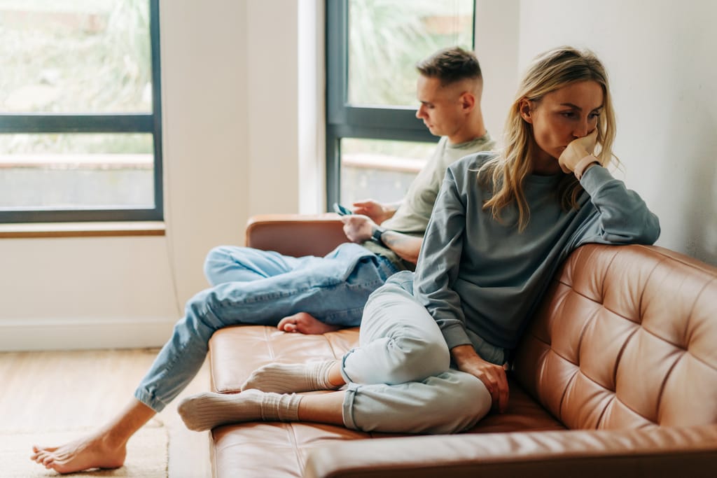 Pariskunta istuu samalla sohvalla fyysisesti lähekkäin mutta emotionaalisesti erillään. Nainen nojaa käsivarteensa ja katsoo alaspäin mietteliäänä, kun mies keskittyy puhelimeensa. Kuvassa korostuu yksipuolinen ja epätasapainoinen suhde.