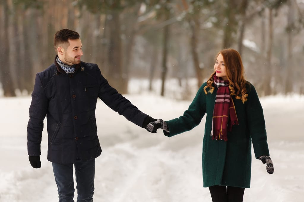 Nuori mies ja nainen kävelevät käsi kädessä lumisessa metsämaisemassa, hymyilevät toisilleen ja ovat pukeutuneet lämpimiin takkeihin, huiveihin ja hansikkaisiin.
