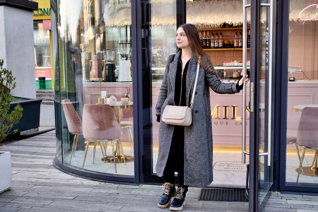 Sama nainen astuu ulos vaaleanpunasävyisestä kahvilasta, pitää lasiovea auki ja katsoo kadulle; hänellä on pitkä harmaa takki, mustat vaatteet, nilkkurit ja beige olkalaukku.