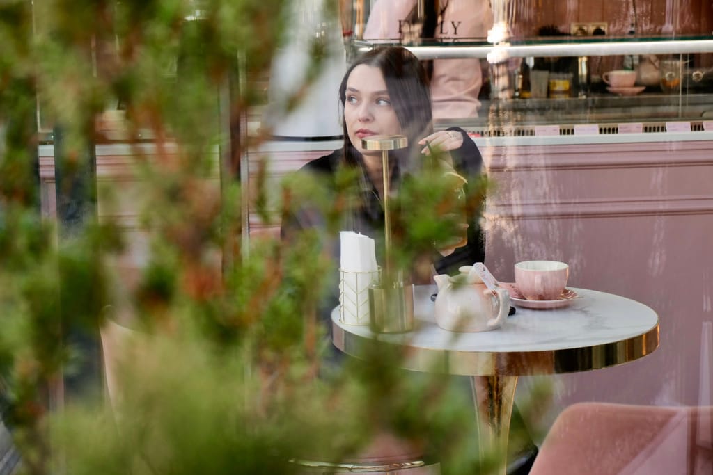 Tummahiuksinen nainen istuu yksin kahvilan pienessä marmoripöydässä ikkunan äärellä; pöydällä on vaalea teekannu, kuppi ja lautanen, ja hän katsoo sivulle mietteliäänä kasvien osittain peittäessä näkymää.