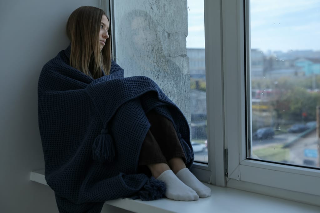 Nuori nainen istuu ikkunalaudalla kääriytyneenä vilttiin ja katsoo ulos mietteliäänä.