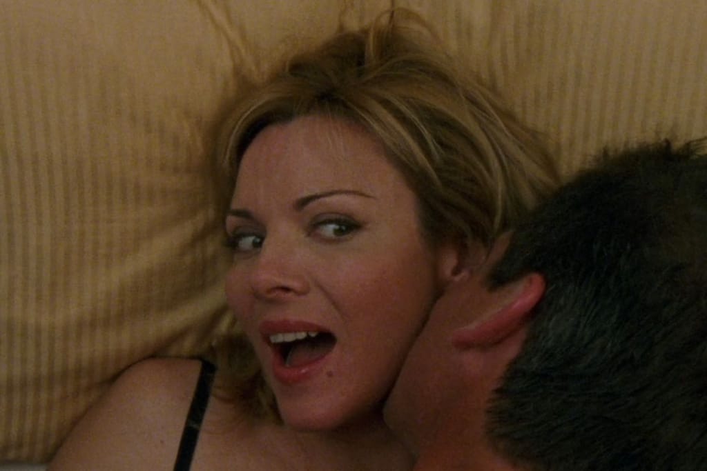 Nainen makaa sängyllä silmät suurina ja mies suutelee hänen kaulaansa. 