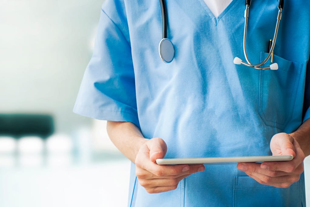 Terveydenhuollon ammattilainen sinisissä työvaatteissa pitää tablettia käsissään, kaulassa stetoskooppi.