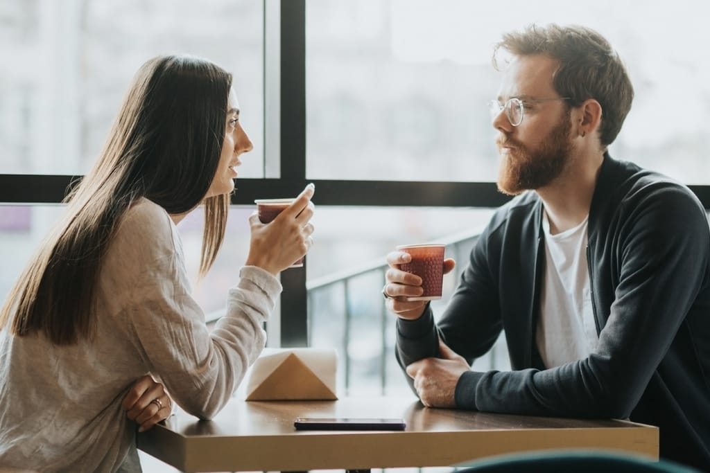 Nainen ja mies istuvat kahvilassa vastakkain ja keskustelevat kahvikupit kädessään.