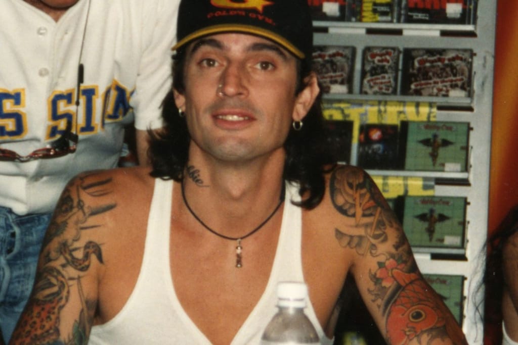 Tatuoitu mies lippalakissa ja valkoisessa hihattomassa paidassa istuu pöydän ääressä ja katsoo kameraan. 