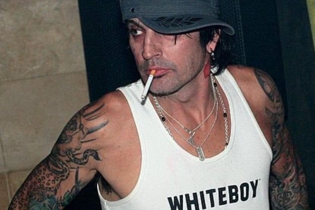 Tatuoitu mies lippalakissa ja valkoisessa hihattomassa paidassa katsoo sivulle savuke suussaan.