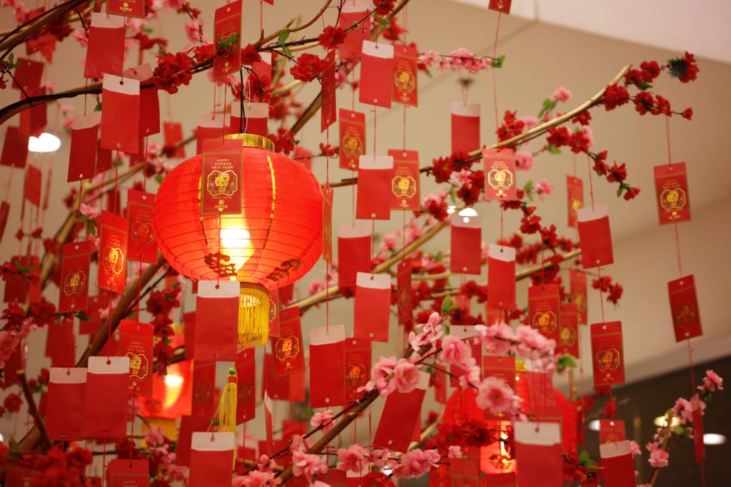 Punainen kiinalainen lyhty ja kukkiva koristepuu, johon on ripustettu punaisia uudenvuoden toivotuskortteja.
