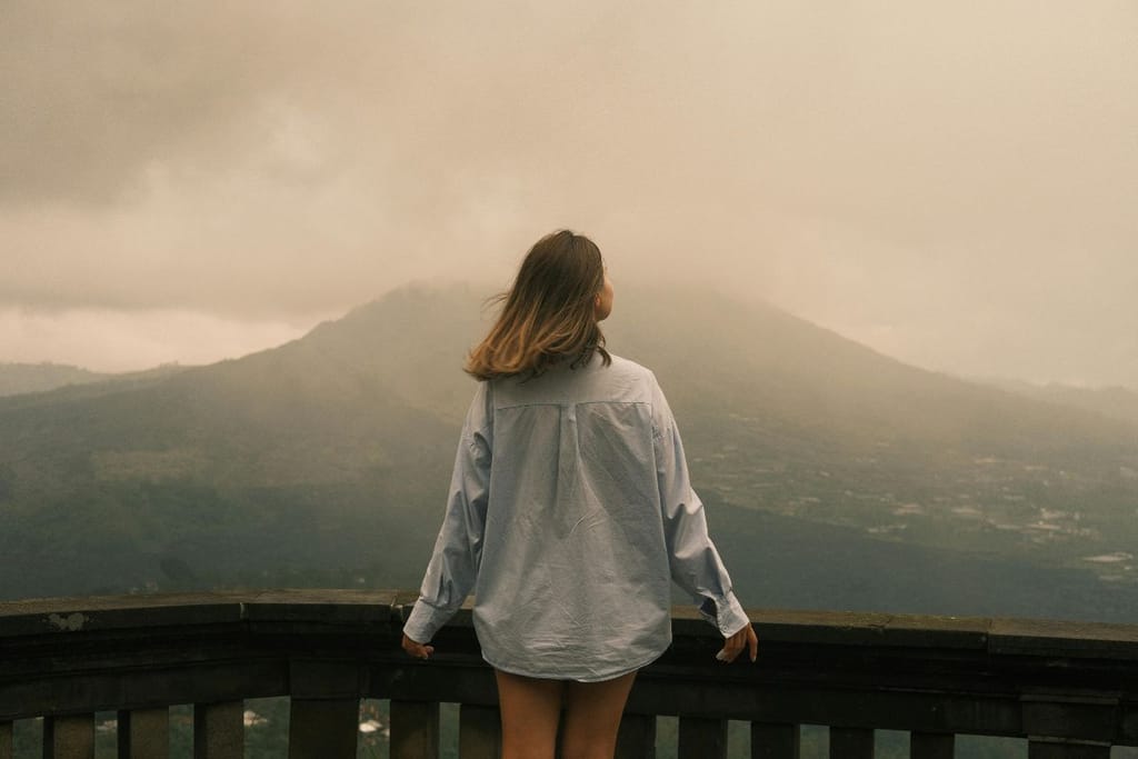Nainen seisoo selkä kameraan päin ja katselee sumuisille vuorille.