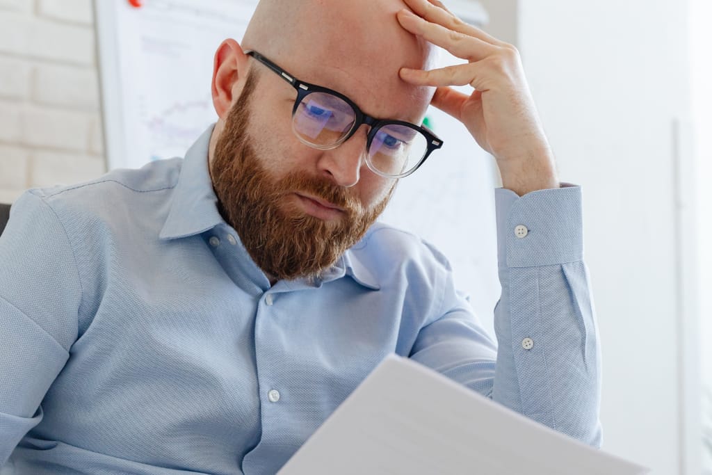 Silmälaseja käyttävä parrakas mies pitelee päätään ja katsoo huolestuneena paperia toimistoympäristössä.
