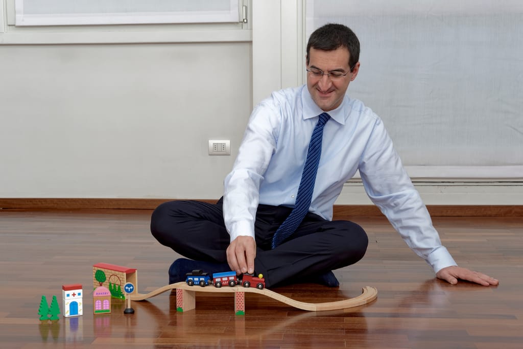 Pukuun ja solmioon pukeutunut mies istuu lattialla ja leikkii puisella junaradalla ja pienillä lelujunilla sisätiloissa.