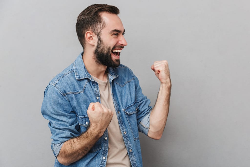 Iloinen parrakas mies nostaa nyrkit ilmaan riemuiten harmaata taustaa vasten.