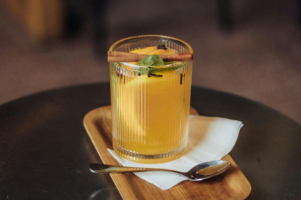 Keltainen juoma lasissa tarjottimella, päällä kanelitanko ja yrtinlehti, asetettuna pöydälle.