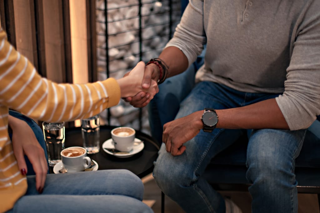 Kaksi ihmistä kättelee toisiaan kahvilassa pienessä pöydässä, jolla on kaksi cappuccinoa ja vesilasit; he istuvat vastakkain rennossa tunnelmassa.