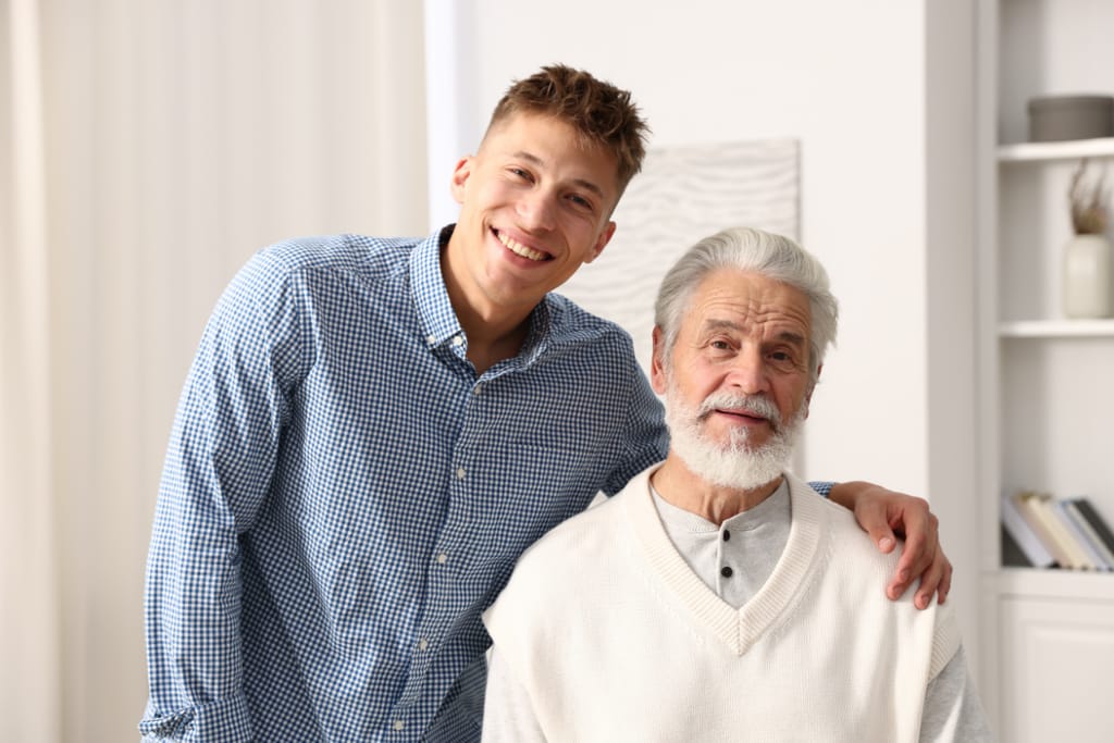 Nuori mies seisoo iäkkään miehen vieressä käsi tämän olkapäällä, molemmat hymyilevät valoisassa sisätilassa.