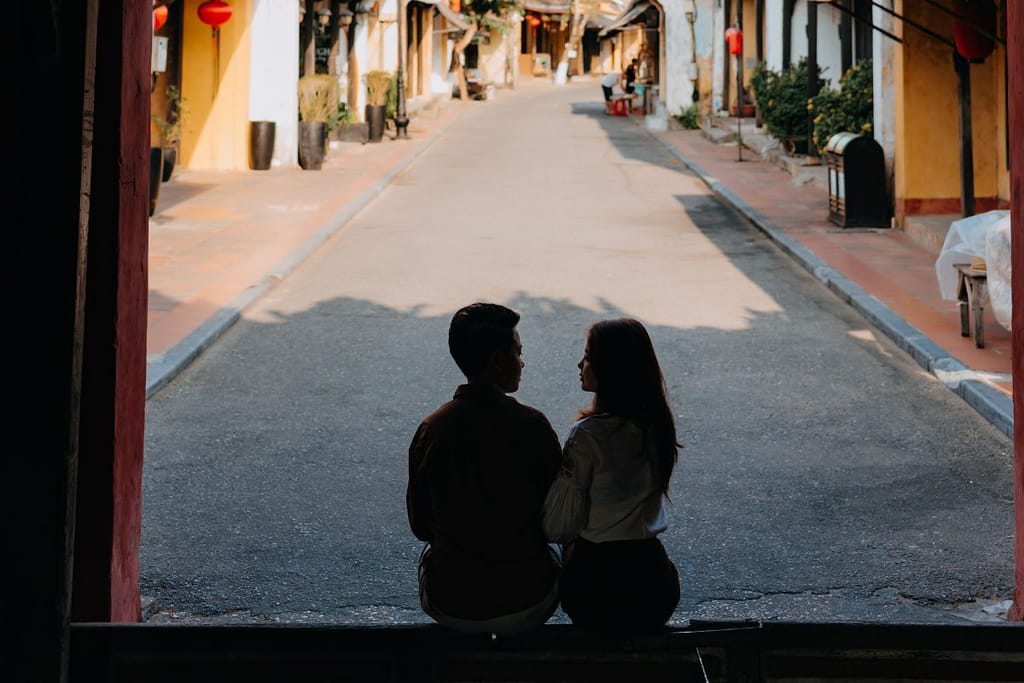 Pariskunta istuu varjossa porttikongin alla ja katsoo toisiaan tyhjällä kadulla.