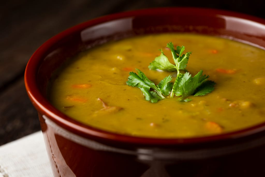 Punaisessa kulhossa tarjoiltu paksu hernekeitto, jonka pinnalla näkyy porkkanapaloja ja yrttejä. Keitto on koristeltu tuoreella persiljalla, ja kulho on asetettu tummalle alustalle.