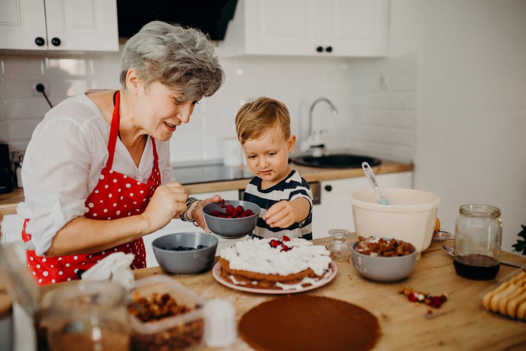 Isoäiti ja pieni poika leipovat yhdessä keittiössä. Poika koristelee kermakakkua marjoilla, ja isoäiti pitää kädessään kulhoa, pöydällä on leivontatarvikkeita ja marjoja.