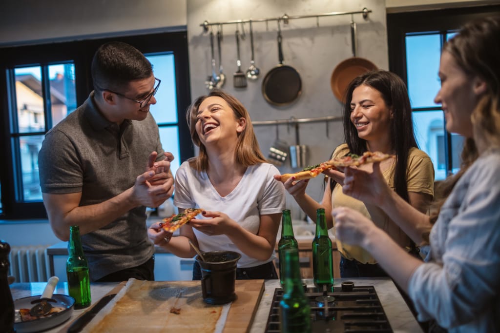 Neljä nuorta aikuista nauraa ja syö pizzapaloja keittiössä; he seisovat lieden ääressä, pöydällä on vihreitä juomapulloja ja taustalla roikkuu paistinpannuja seinällä.