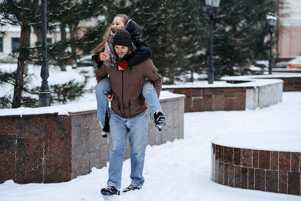 Nuori mies kantaa naista selässään lumisessa kaupunkiympäristössä. Molemmat ovat pukeutuneet talvivaatteisiin, ja nainen nauraa miehen selässä heidän kulkiessaan lumisella kävelytiellä puiden ja kivirakenteiden vieressä.