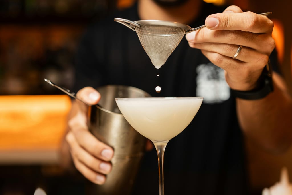 Baarimikko siivilöi vaaleaa cocktailia lasiin metallisen siivilän avulla.