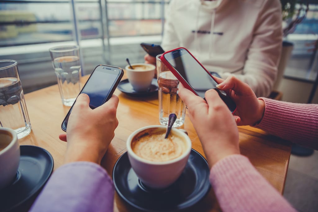 Usean henkilön kädet pitelevät älypuhelimia kahvilapöydän ääressä, pöydällä kahvikuppeja ja vesilaseja