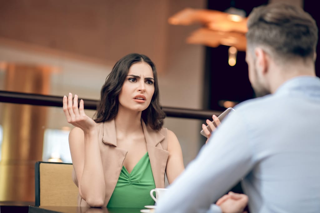 Nainen ja mies istuvat pöydän ääressä sisätiloissa, nainen elehtii kädellään ja pitää älypuhelinta, pöydällä on kahvikuppi