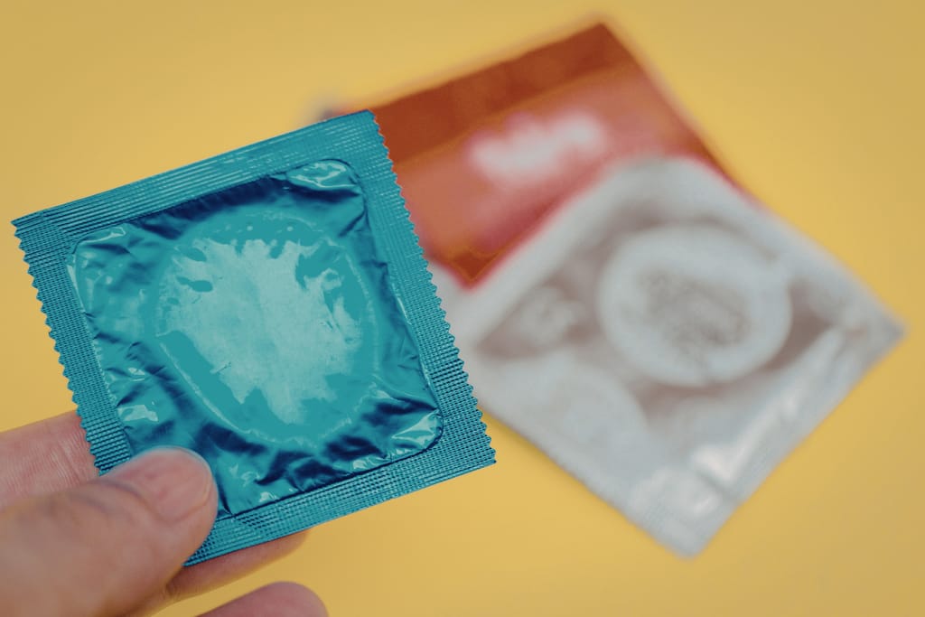 Käsi pitelee sinistä kondomipakkausta, taustalla toinen avoin pakkaus, jossa näkyy rullattuja kondomeja keltaista taustaa vasten.