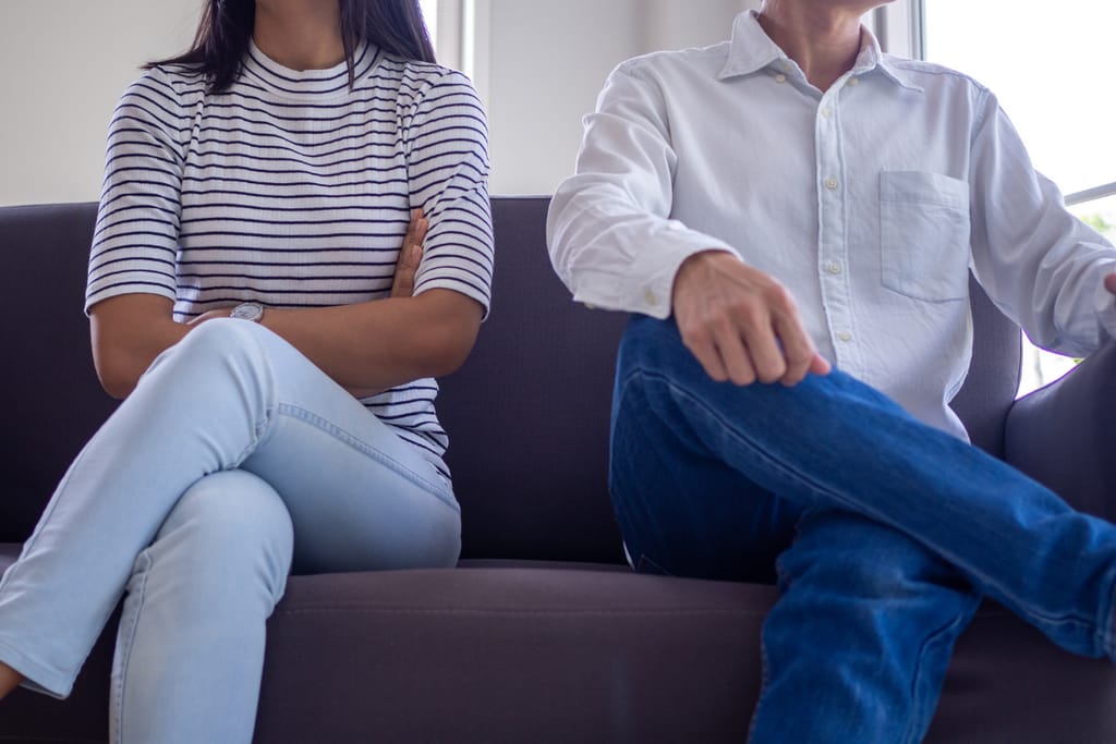 Kaksi aikuista istuu sohvalla vierekkäin, molemmilla jalat ristissä ja kädet vartalon edessä