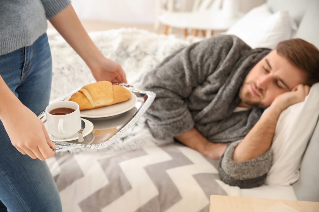 Henkilö tuo tarjottimella kahvia ja croissantin sängyssä makaavalle miehelle, joka lepää kyljellään peiton alla.