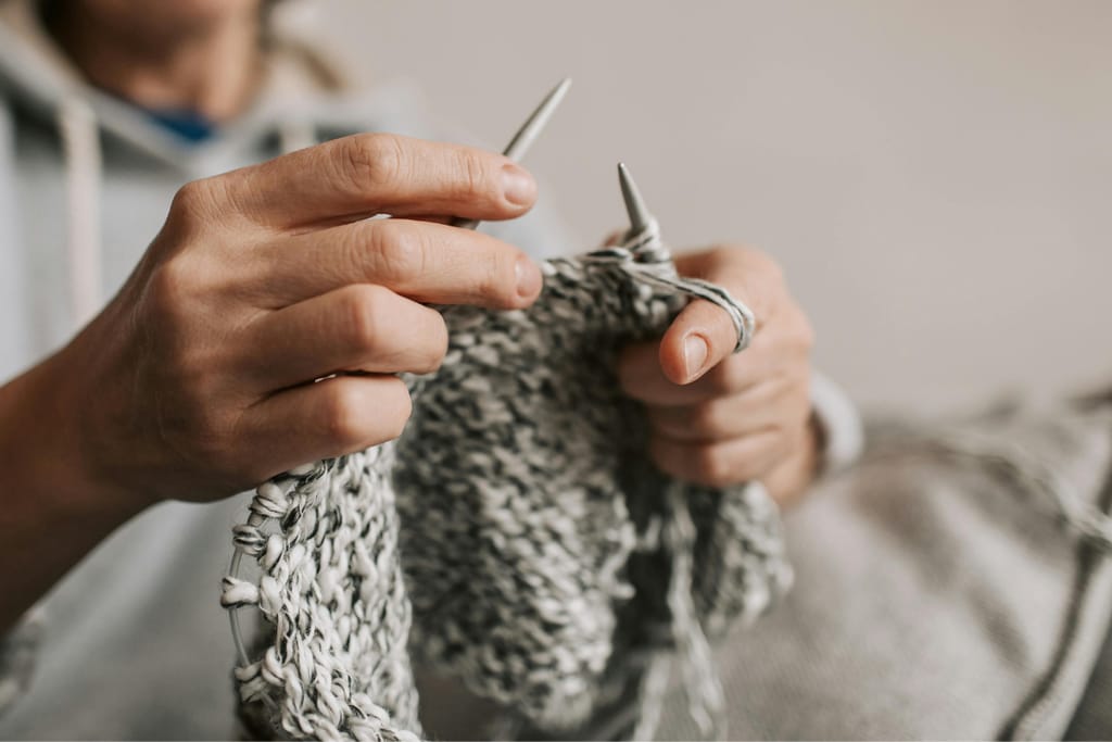 Lähikuva käsistä, jotka neulovat harmaasävyistä lankaa puikoilla.