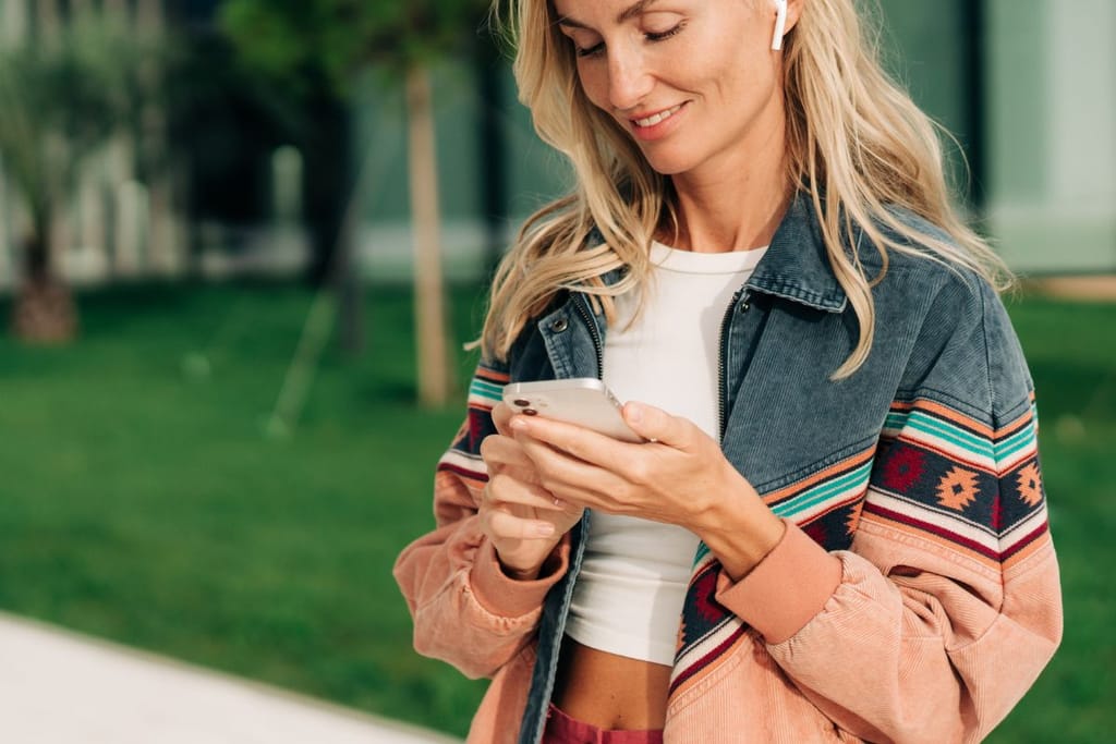 Nainen seisoo ulkona modernin rakennuksen edessä ja katsoo puhelintaan, korvissaan langattomat kuulokkeet.