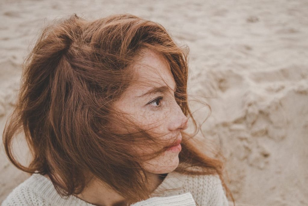 Punatukkainen nainen katsoo sivulle tuulisella rannalla hiukset kasvoilla.