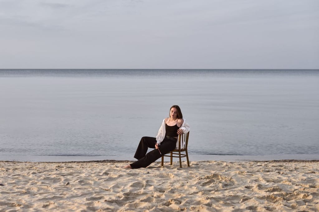 Nainen istuu rannalla puutuolilla yksin ja hän pitelee kädessään sivellintä.