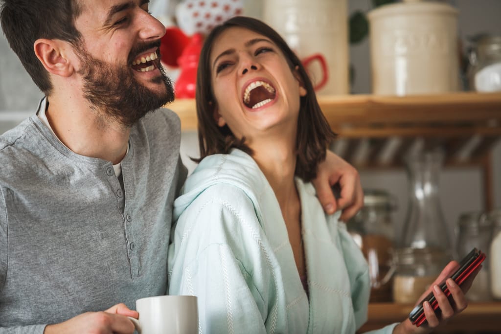 Iloinen pariskunta nauraa keittiössä lähekkäin, miehellä kädessään muki ja naisella puhelin.