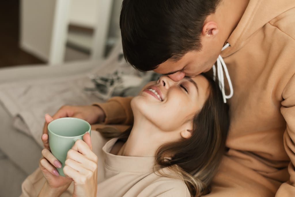 Pariskunta rentoutuu sohvalla; mies suukottaa hymyilevää naista otsalle, ja nainen pitelee kädessään vihreää kahvikuppia.