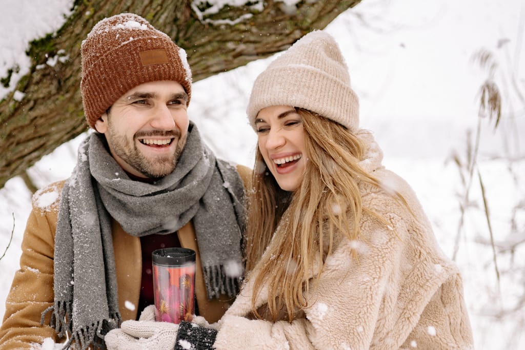 Iloinen pariskunta nauraa lumisessa metsässä, toisella kädessä termospullo ja molemmilla talvivaatteet.