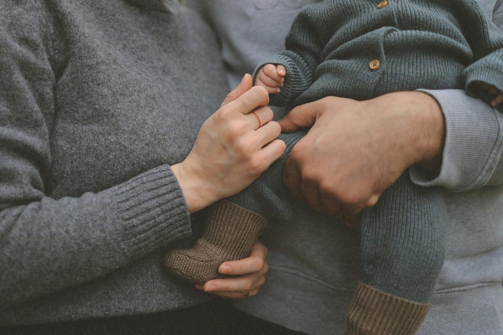 Aikuisten kädet pitävät vauvaa sylissä, ja toinen vanhemmista koskettaa hellästi vauvan pientä kättä.