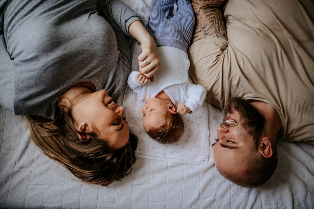 Äiti ja isä makaavat sängyllä vastakkain vastasyntyneen vauvan kanssa ja hymyilevät lapselleen.
