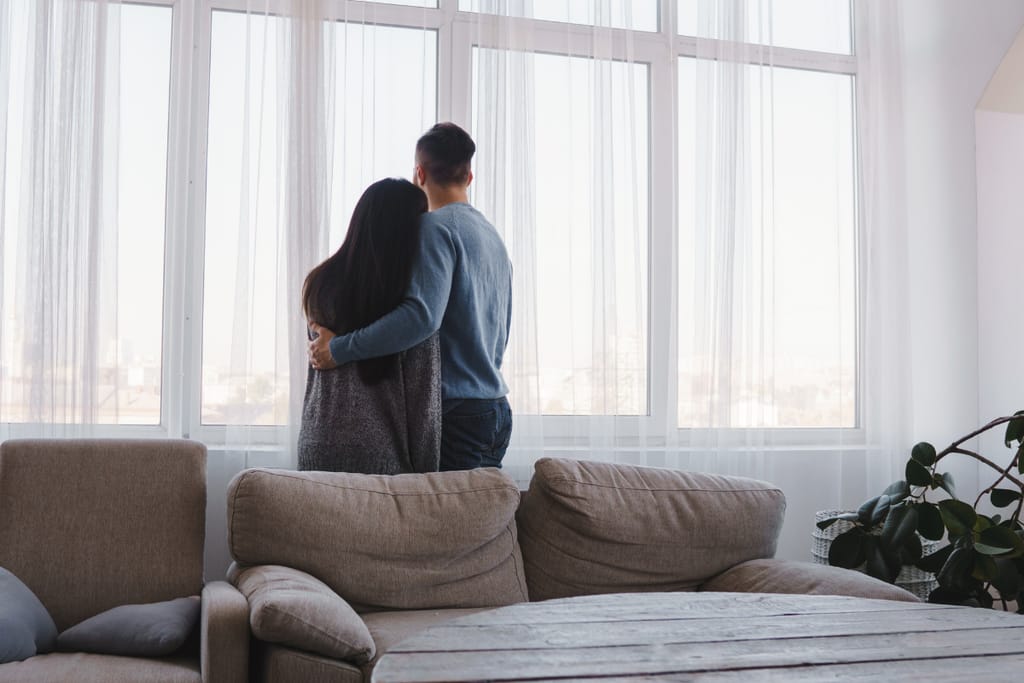 Pariskunta seisoo vierekkäin olohuoneessa suuren ikkunan edessä, mies pitää kättään naisen hartioilla ja he katsovat ulos valoisaan maisemaan. Etualalla näkyy beige sohva ja puinen sohvapöytä, oikealla viherkasvi.