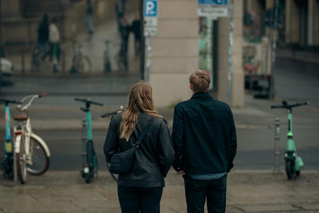 Kaksi aikuista henkilöä kävelee selin kadulla, sähköpotkulautoja ja polkupyöriä ympärillä