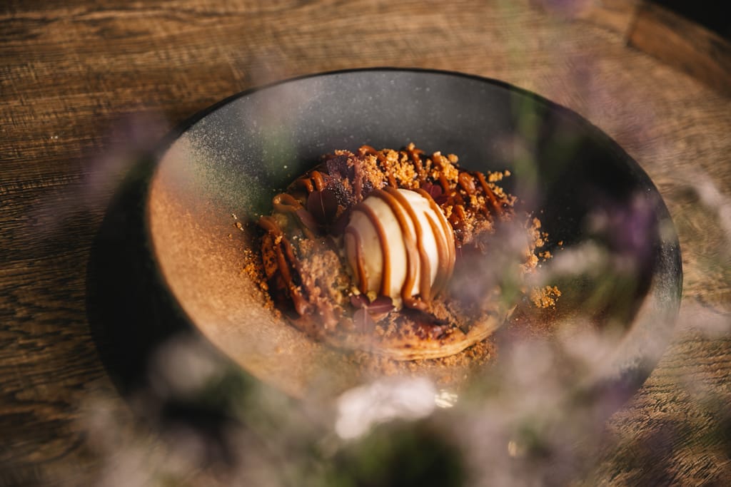 Jälkiruoka mustassa kulhossa: vaniljajäätelöpallo, jonka päällä kinuskikastiketta, suklaata ja murua puupöydällä.
