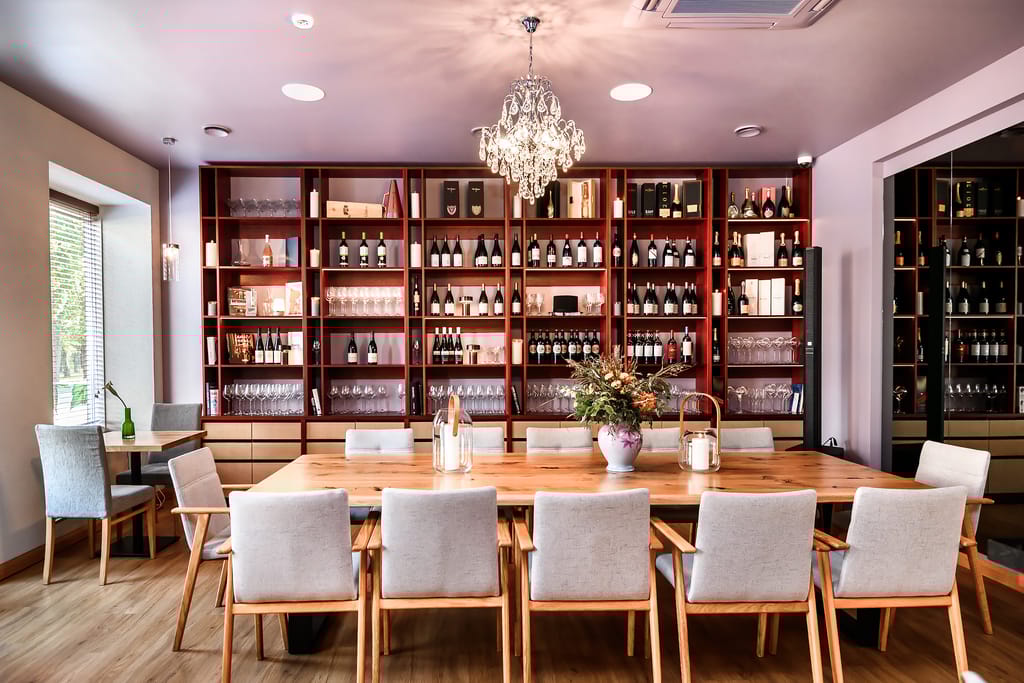 Ravintolan tyylikäs sisätila: pitkä puinen pöytä, vaaleat tuolit ja seinällinen viinipulloja hyllyissä.