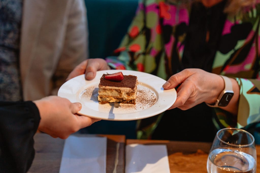Jälkiruoka-annos ojennetaan pöydässä: pala kerroksellista kakkua valkoisella lautasella.
